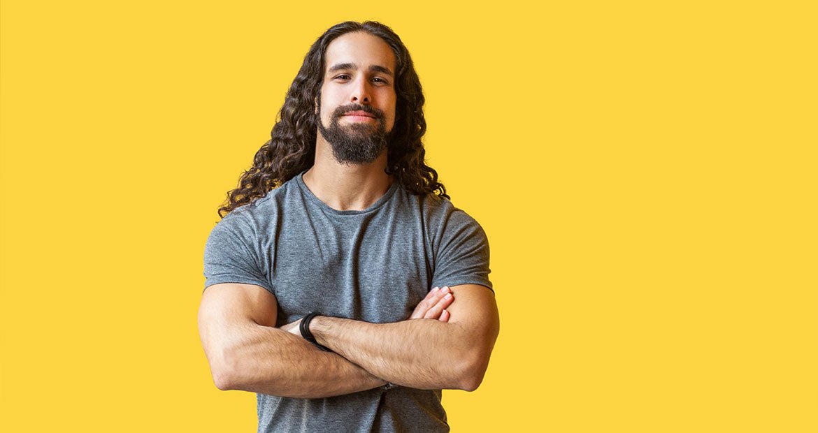 Homem com cabelo masculino grande cruza os braços enquanto sorri levemente